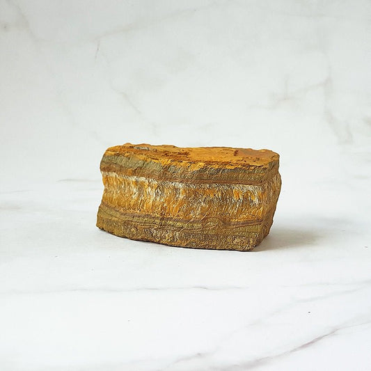 Ángulo de un ojo de tigre en bruto con vetas doradas y marrones, mostrando su textura natural y forma irregular.